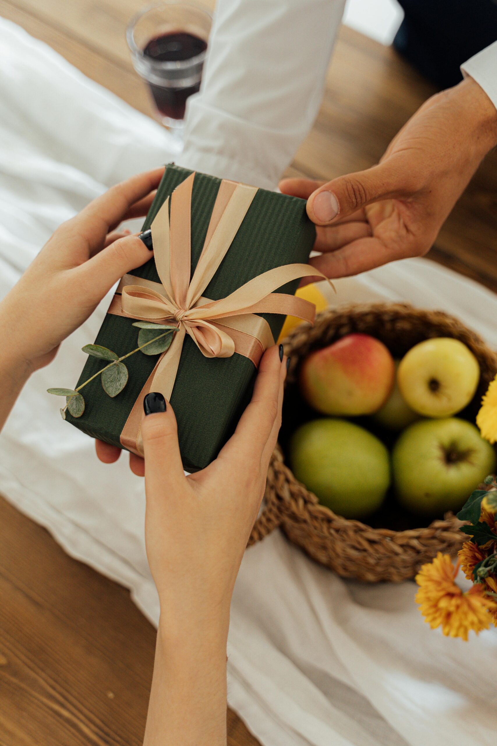 A close-up of hands exchanging a green gift next to a fruit basket on a wooden table.